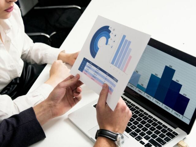 Hands hold a sheet of charts in front of a computer screen with a bar chart on it for our post on digital marketing consultants. (Photo Credit: Artem Podrez).