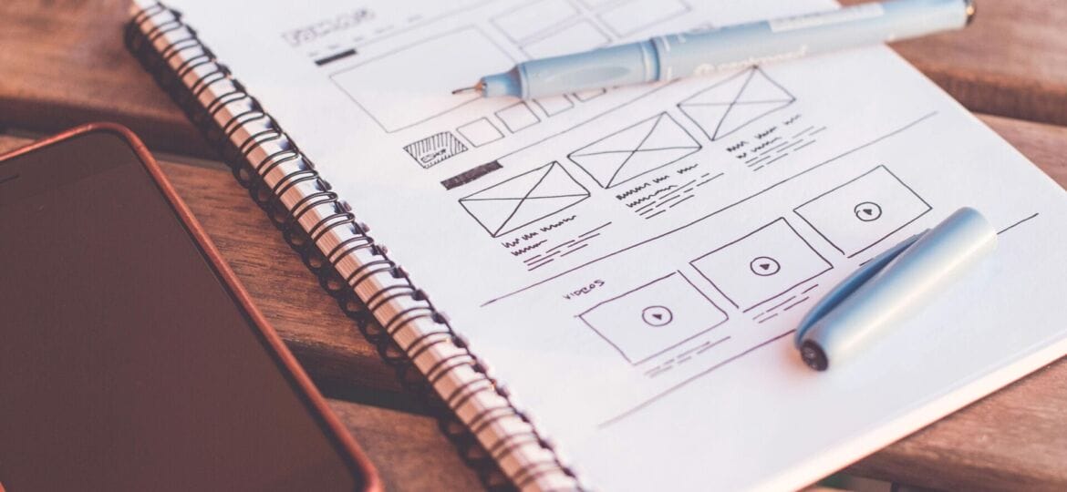 A phone and a wireframe skectch in a journal sit on a wooden table.