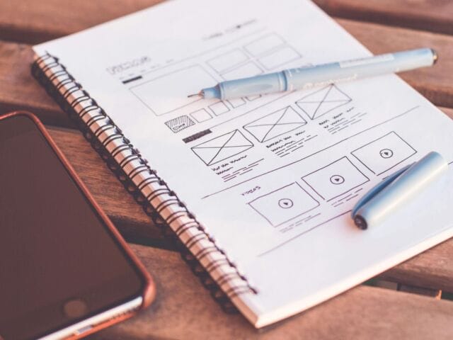A phone and a wireframe skectch in a journal sit on a wooden table.